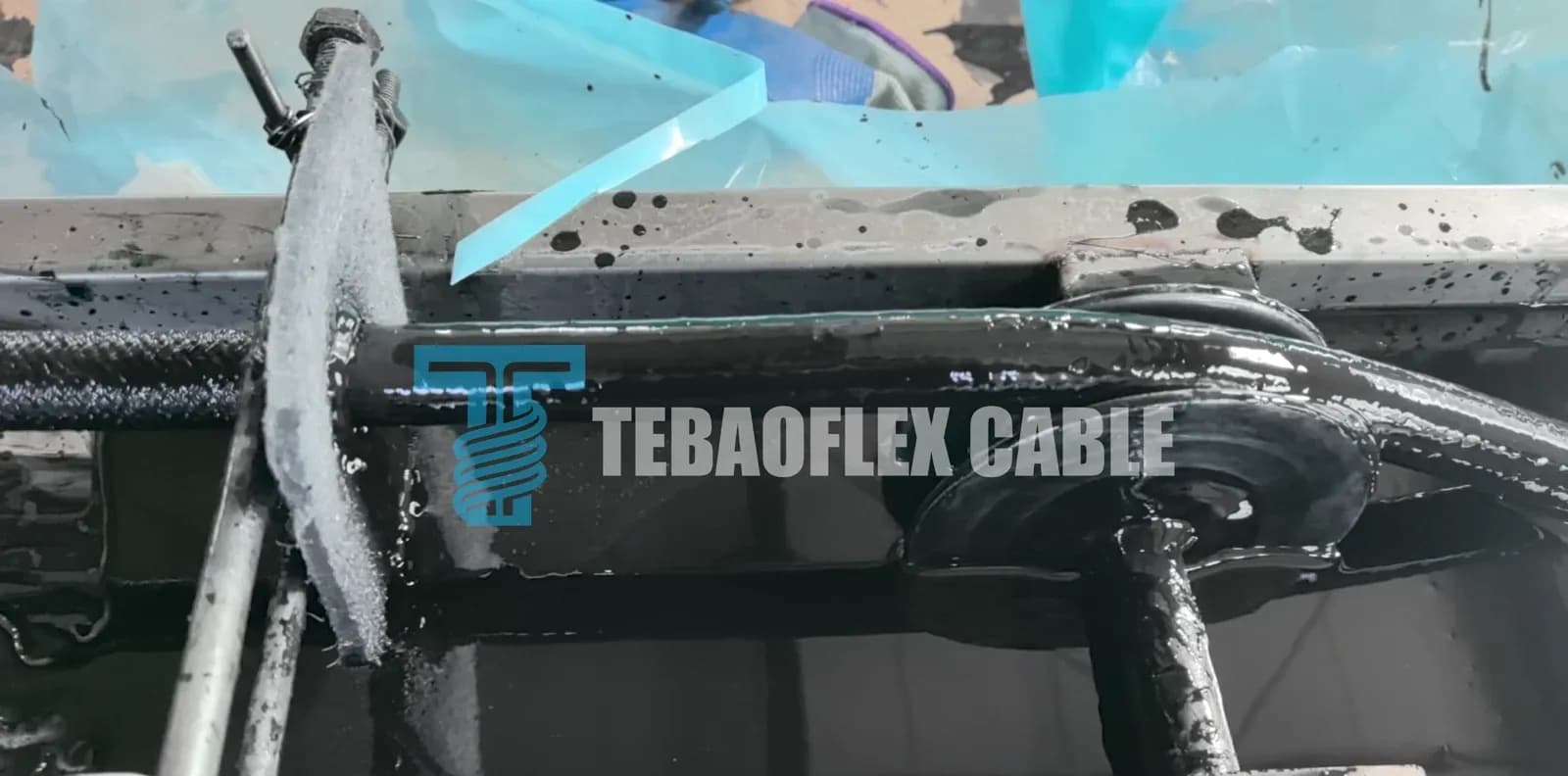 A braided cable running through an open trough of dark fluid, with a fibrous pad clamped against it by a threaded adjuster; the cable surface is glossy and wet on one side of the pad and matte with visible braid texture on the other.