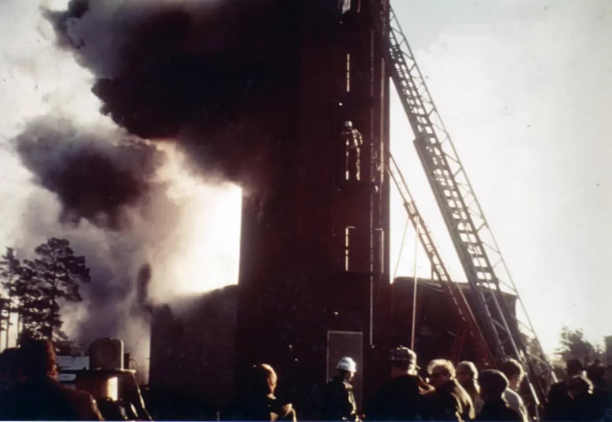 Black smoke pouring from a small fire-test building, with a fire-brigade ladder and onlookers in the foreground