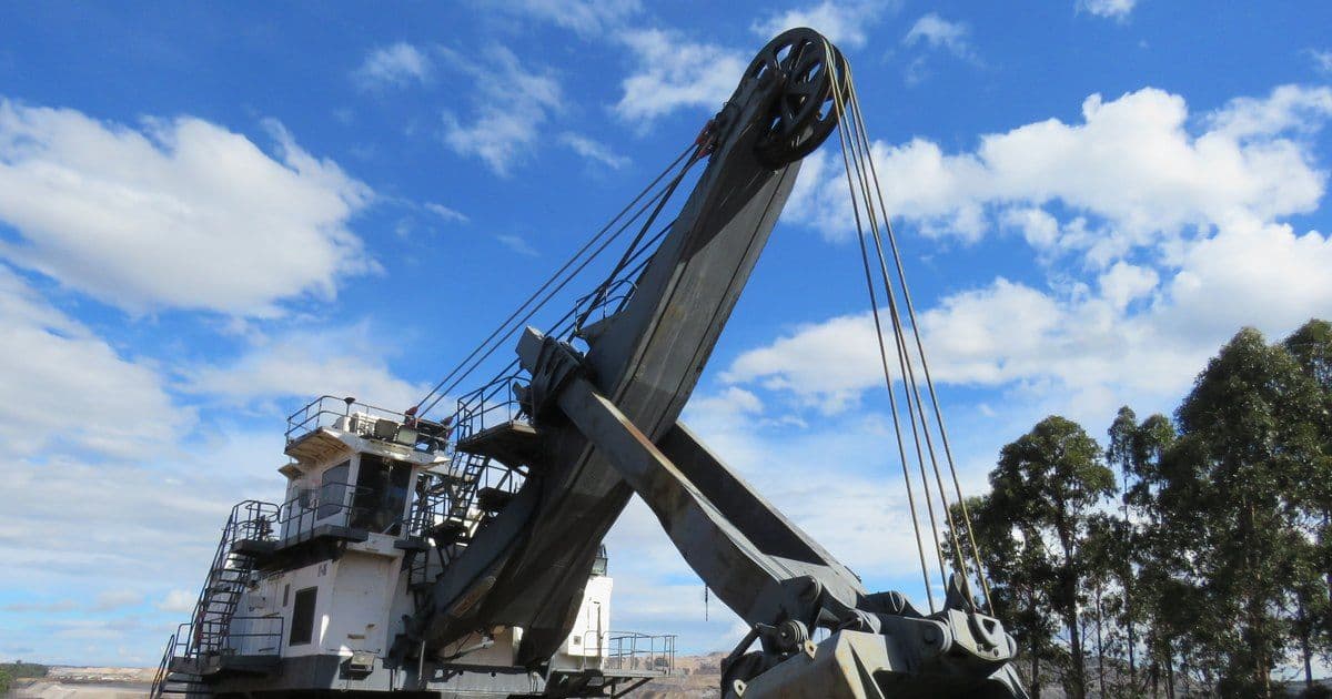 A retired electric rope shovel with a tall boom, hoist ropes and a large dipper, on display at the edge of an open-cut coal mine under a blue sky