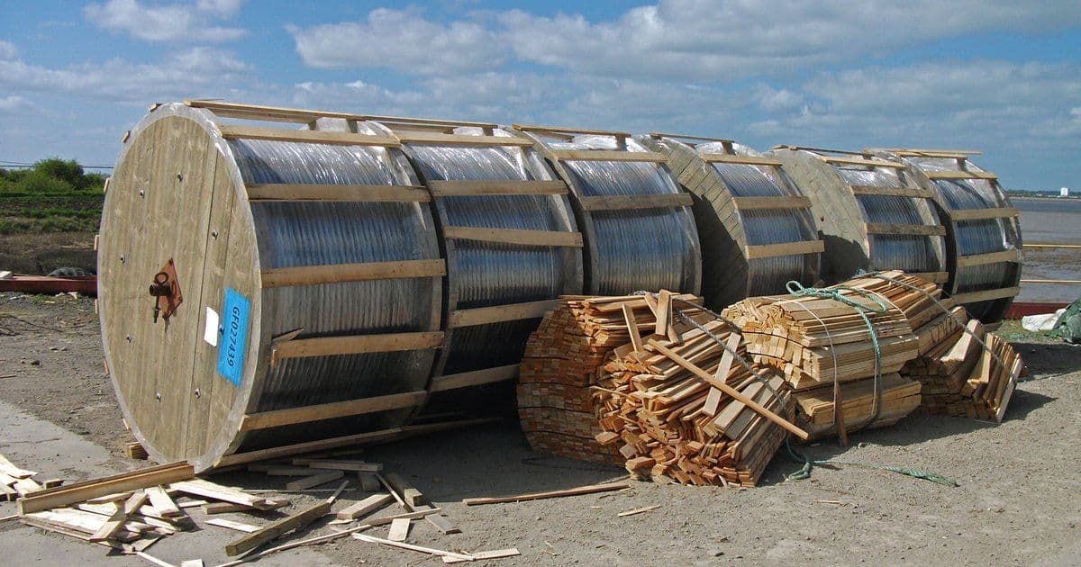 Large cable drums wrapped in plastic and closed with wooden battens, lying on a quayside with bundles of loose battens beside them