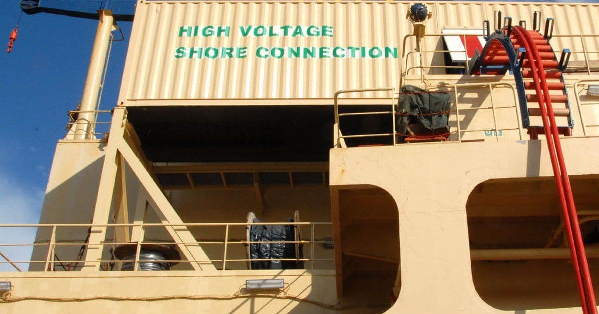 Red power cables run down a ship's side from a deck frame beside a container marked High Voltage Shore Connection