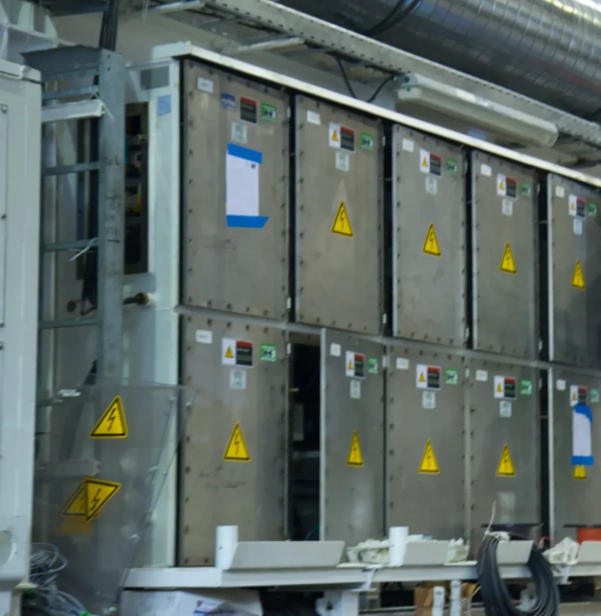 Grey electrical cabinets with yellow high-voltage warning signs under a ventilation duct on a tunnel boring machine gantry