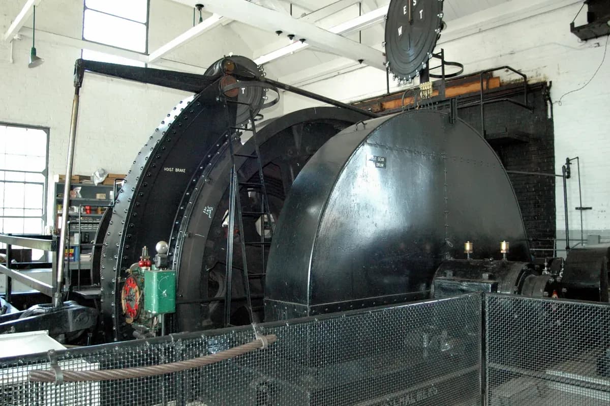 A large black mine hoist drum and brake inside a brick engine house, behind a wire-mesh guard