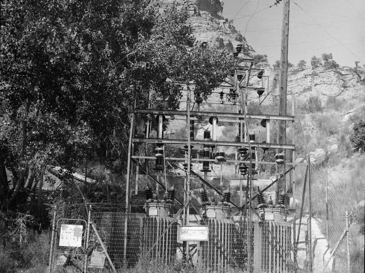 A fenced coal mine substation with three transformers under a steel frame of insulators, below a rocky hillside